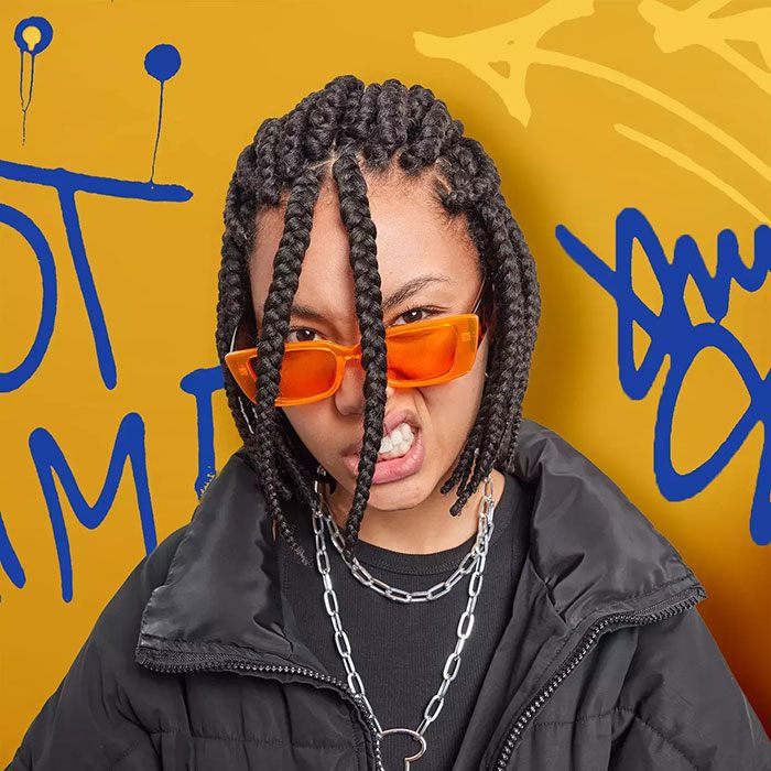 Person mit geflochtenem Haar, schwarzer Jacke und silberner Kette vor gelbem Hintergrund mit blauer Graffiti-Schrift