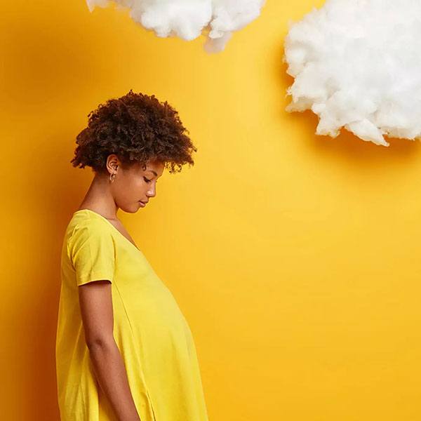 Schwangere Frau in gelbem Kleid vor gelber Wand mit zwei weißen Wolken aus Watte oder Stoff