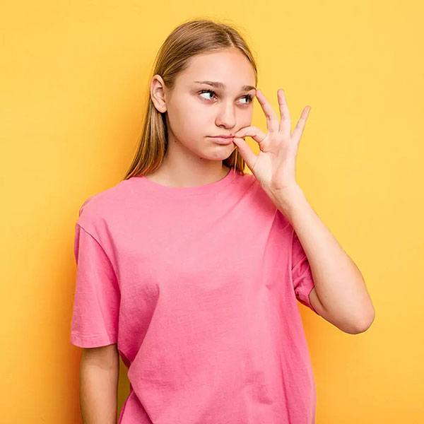Junge Frau mit langen Haaren trägt ein pinkes T-Shirt und zeigt mit der rechten Hand das Verschwiegenheitszeichen vor einem gelben Hintergrund