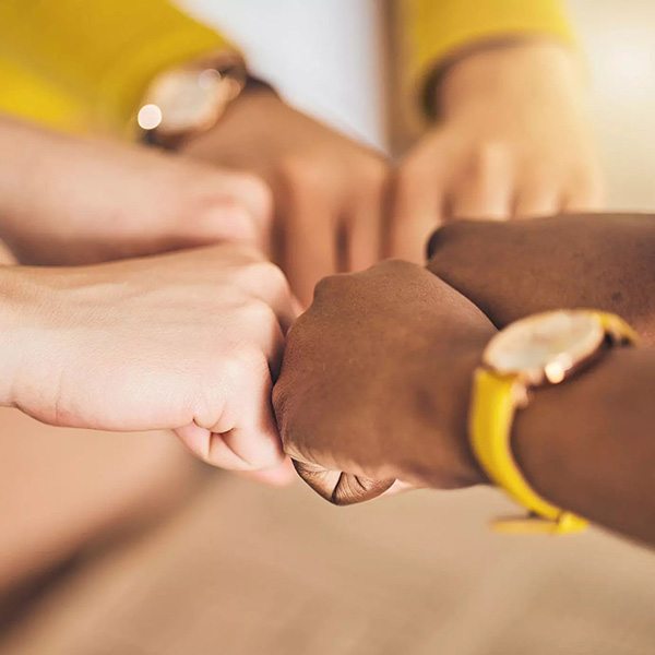 Vier Fäuste stoßen sich in der Bildmitte zusammen, zwei Personen tragen gelbe Armbanduhren